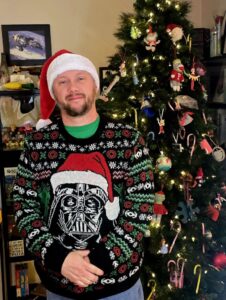 chris cox in a festive outfit standing next to a decorated christmas tree.